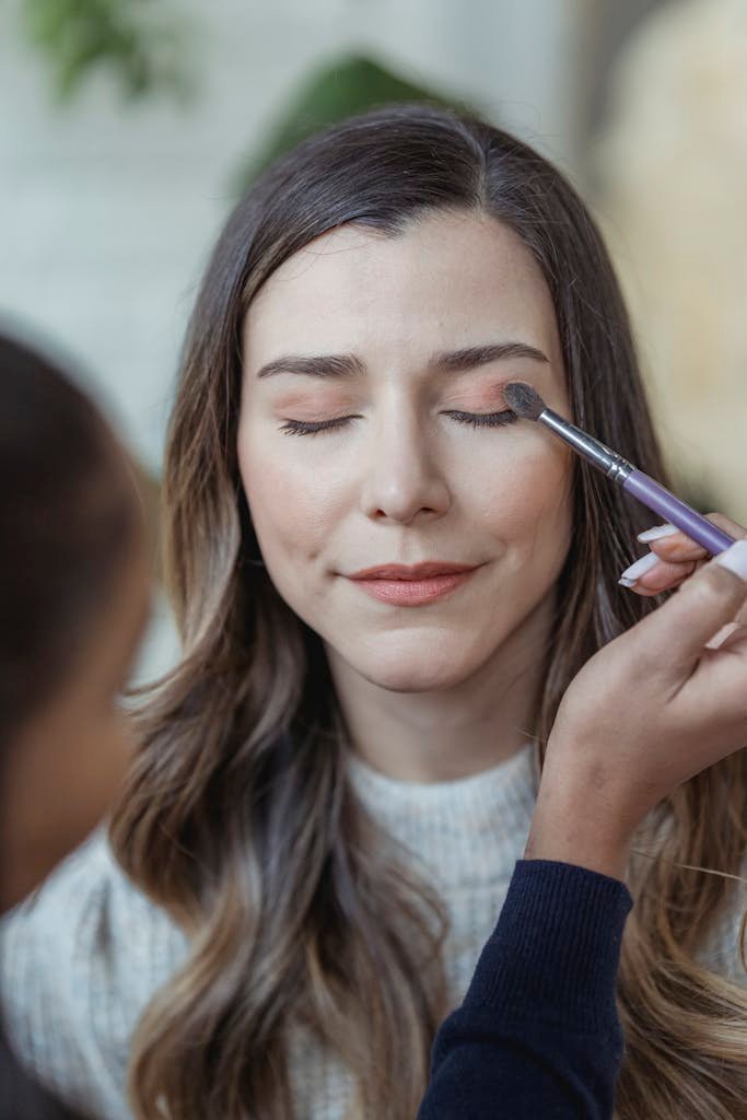 Makeup artist with brush applying eyeshadows on glad female customer in light beauty studio