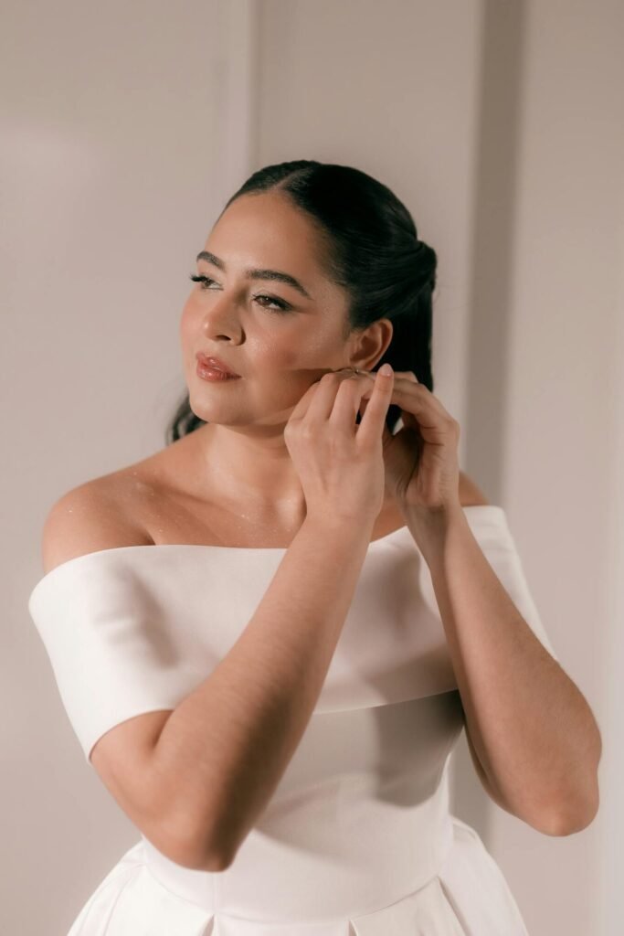 A bride elegantly adjusts her earring, dressed in a stylish white gown indoors.