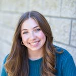 A cheerful woman in a teal sweater sits against a brick wall, beaming with joy. Captured outdoors, showcasing natural beauty.