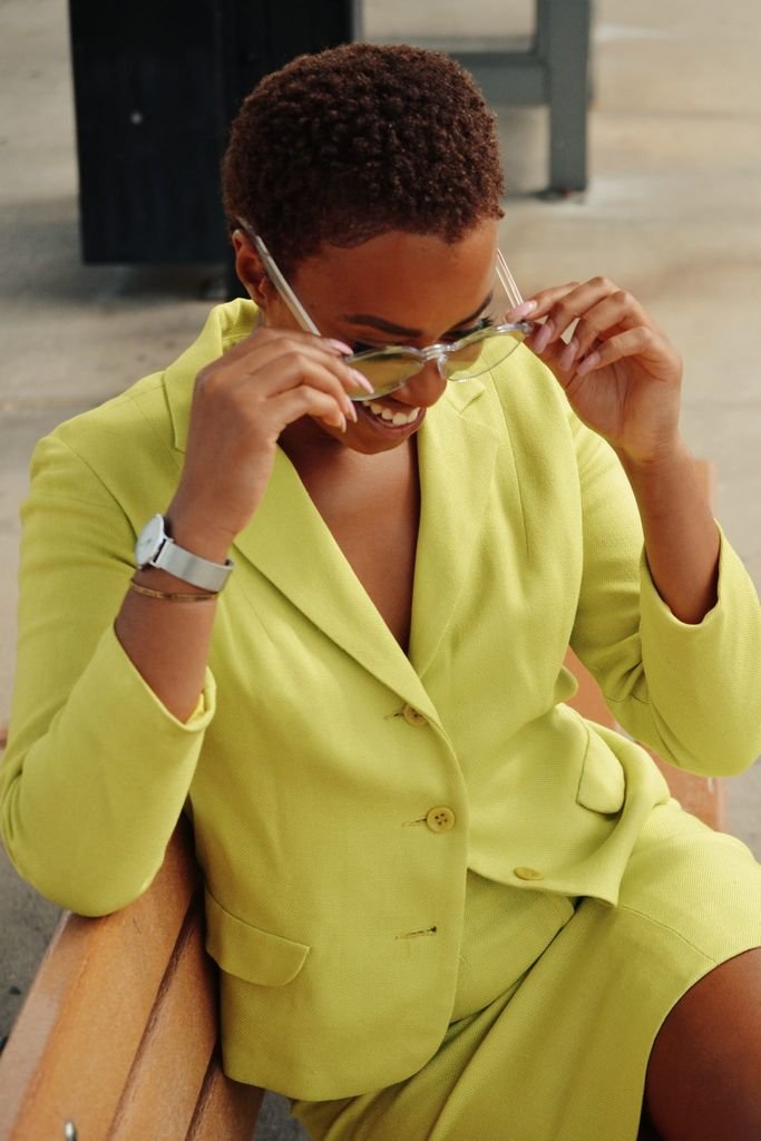A fashionable woman in a bright yellow suit adjusts her glasses while sitting outdoors on a sunny day.