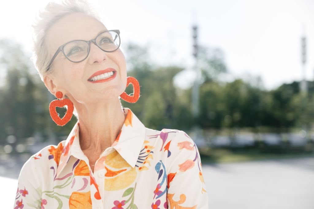Charming senior woman with glasses and heart earrings smiles joyfully outdoors in floral attire.