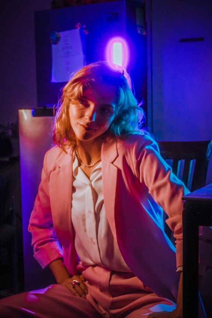 Elegantly posed woman in a pink suit under vibrant neon lighting in a moody setting.